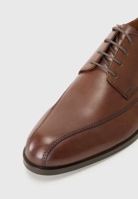 Chaussure habillée en cuir marron avec un bout pointu, ornée de coutures tonales et de lacets gris. Texture lisse et design épuré.