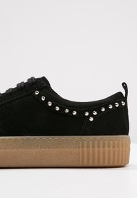 Baskets en cuir suédé noir avec des clous argentés le long du col et du talon, semelle épaisse en caoutchouc marron clair texturée, et lacets noirs.