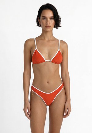 Donna con capelli corti e scuri che indossa un bikini arancione con bordino bianco, in piedi davanti a uno sfondo bianco semplice, rivolta in avanti.