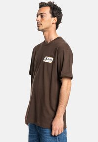 Brown cotton T-shirt with a round neckline and short sleeves, featuring a white and yellow branded patch on the left chest.