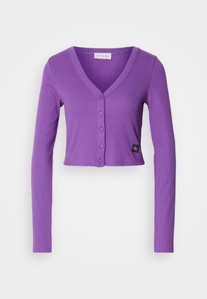 Purple cropped cardigan with long sleeves, a V-neck, and a button-down front. Features ribbed texture and a branded tag on the hem.