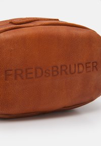 Bruine leren tas met een getextureerd oppervlak, met aan de zijkant het geperste merk "FREDsBRUDER". Ronde vorm met een gladde afwerking.