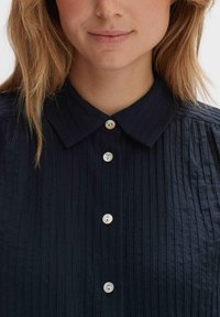 Navy blue shirt with a textured, pleated design, featuring a pointed collar and shiny, rounded buttons along the front.