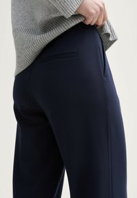 Pantalon marine en tissu lisse, avec une taille plate, une poche arrière discrète et une coupe ajustée, associé à un pull en maille gris clair.