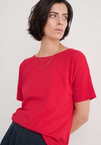 Rotes T-Shirt mit kurzen Ärmeln, aus weichem Stoff und lockerem Schnitt, mit einem runden Ausschnitt, getragen über schwarzen Hosen. Sichtbare goldene Kettenhalskette.