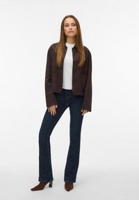 Žena stoji noseći tamnosmeđi sako na dugmad, bijelu majicu, tamnoplave traperice i zakovike smeđe cipele s visokom petom na bijeloj pozadini.