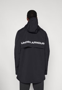 Μαύρο αδιάβροχο μπουφάν με κουκούλα, με εμφανές λευκό λογότυπο "UNDER ARMOUR" στην πλάτη και κομψό σχεδιασμό.