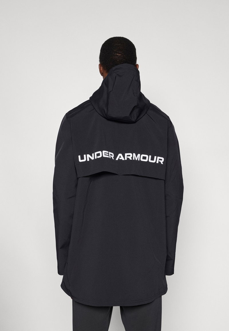 Μαύρο αδιάβροχο μπουφάν με κουκούλα, με εμφανές λευκό λογότυπο "UNDER ARMOUR" στην πλάτη και κομψό σχεδιασμό.