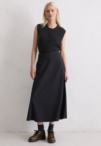 Haut sans manches noir avec un tricot texturé et des boutons, associé à une longue jupe noire. Des chaussures à lacets marron et des chaussettes foncées complètent le look.