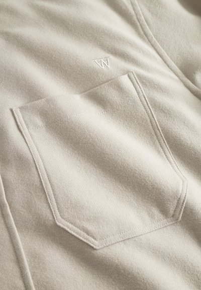 Tissu de chemise beige avec une texture visible, doté d'une grande poche à rabat et d'un petit logo blanc brodé sur le côté.
