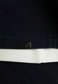 Navy kangasta, jossa on sileä pinta, valkoisella joustavalla vyötärönauhalla ja mustalla kolmiomaisella tarralapulla, jossa on minimalistinen valkoinen muotoilu.