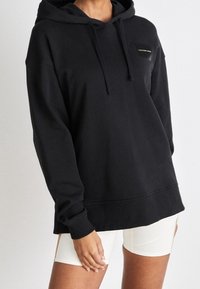 Personer i en svart Calvin Klein hoodie med dragsko och vita cykelshorts, poserar mot en enfärgad ljus bakgrund.