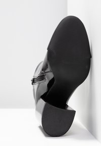 Botte en cuir noir brillant inclinée contre un coin blanc, montrant une semelle lisse et un talon bloc texturé.