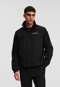 Schwarze wasserdichte Jacke mit hohem Kragen, Druckknöpfen und Fronttaschen. Logo auf der Brust, mit elastischen Bündchen und Saum.