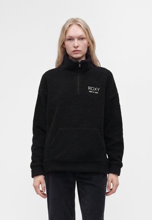 Femme aux longs cheveux blonds portant un pull polaire noir avec le logo "ROXY SINCE 1990", debout devant un fond blanc uni.
