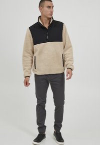 Beige fleece pullover with a black nylon quarter-zip collar, side pockets, and elastic cuffs, paired with gray pants and black sneakers.