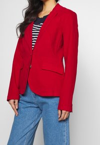 Červený blazer se skvělým střihem, s jednobotonovým zapínáním a dvěma předními kapsami. Nesený přes námořnický pruhovaný top a doplněný modrými džíny.