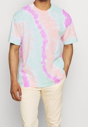 Homme portant un t-shirt tie-dye pastel aux couleurs rose, bleu et beige, associé à un pantalon de couleur crème, debout avec une main dans la poche.