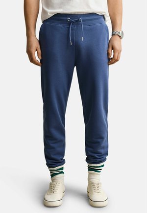 Man draagt blauwe joggers met koord, witte sneakers en witte sokken met groene strepen, staand tegen een witte achtergrond.