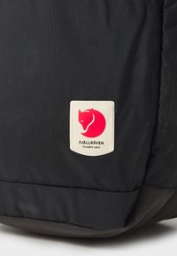 Schwarzer Stoffrucksack mit einem rechteckigen, weißen Patch, auf dem ein rotes Fuchslogo und der Text "FJÄLLRÄVEN Schweden 1960" am unteren Rand zu sehen sind.