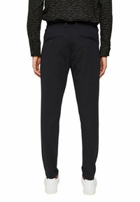 Muž v černých slim-fit kalhotách s kapsami na zadní straně a bílými teniskami, stojící proti jednoduchému bílému pozadí.