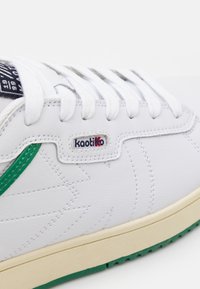 Sneaker in pelle bianca con una striscia di accento verde, punta arrotondata, lacci bianchi, superficie testurizzata e un piccolo etichetta con il marchio sul lato.