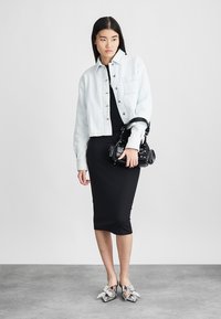 Ανοιχτό μπλε τζιν cropped μπουφάν πάνω από ένα μαύρο εφαρμοστό φόρεμα, συνδυασμένο με μαύρη τσάντα χειρός και ασημένια παντόφλα με διακοσμητικά φιόγκο. Γκρι φόντο.