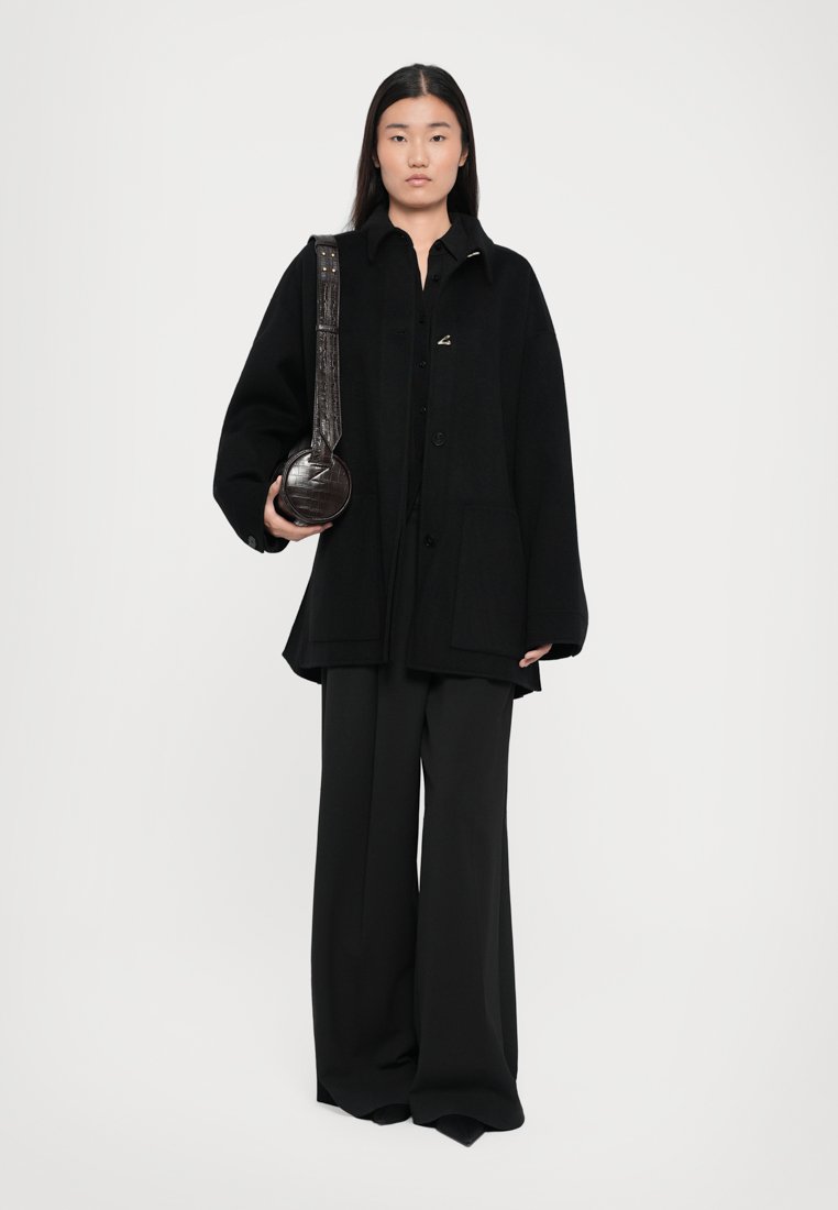 Veste oversize noire, chemise noire, pantalon large noir et un sac à main rond avec une sangle texturée, le tout dans une palette monochromatique.