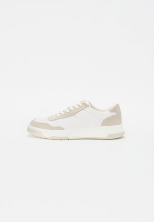 Zapatilla blanca de caña baja con detalles en ante beige, cordones blancos y una suela blanca gruesa sobre un fondo blanco liso.