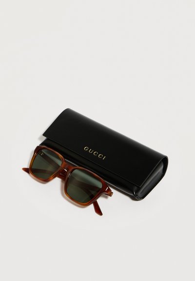Lunettes de soleil à monture carrée marron avec des verres foncés, posées à côté d'un étui Gucci noir avec des inscriptions dorées sur un fond blanc.