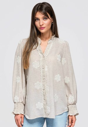 Femme aux longs cheveux bruns porte un chemisier beige avec des fleurs blanches brodées, un col à volants et des poignets froncés, associé à un jean bleu clair.