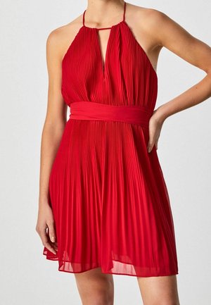 Femme portant une robe rouge plissée sans manches avec un col licou à fente clé et une large ceinture, main reposant sur sa hanche.