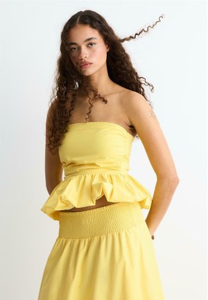 Mladá žena s dlhými kučeravými vlasmi, nosiaca žltý top bez ramienok s volánmi a zodpovedajúcu žltú sukňu, stojaca pred jednoduchým pozadím.