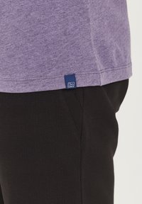 Lilla heather t-shirt med en flad kant, der har et lille blåt logo-tag; stylet med teksturerede sorte bukser.