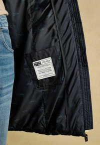 Melnas jakas iekšpuse, kurā redzams Joules zīmola marķējums ar Lielbritānijas karogu, teksts par amatniecību un zilu lapsu rakstu odere, blakus cilvēkam zilos džinsos.