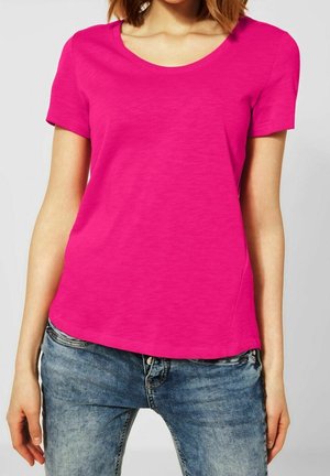 Frau trägt ein knallpinker kurzärmeliger T-Shirt und blaue Jeans, steht mit entspannten Armen an den Seiten.