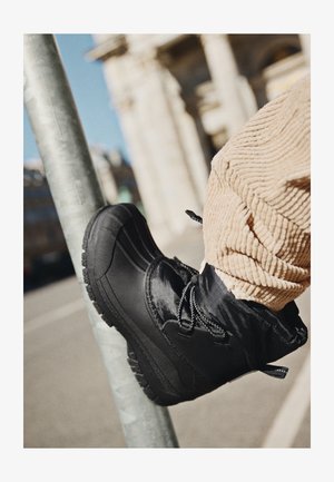 Bota negra de invierno con suela gruesa, usada con pantalones de pana beige apoyada en un poste metálico en un entorno urbano al aire libre.
