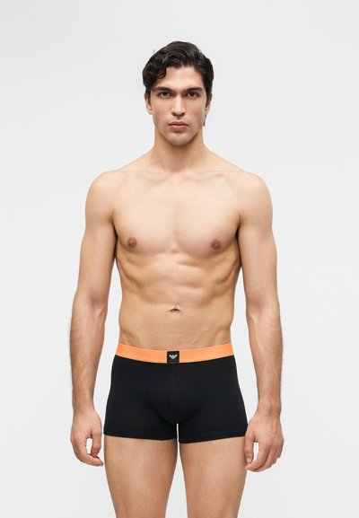 Junger Mann mit dunklen Haaren, der nach vorne blickt und schwarze Boxershorts mit einem orangefarbenen Bund vor einem schlichten weißen Hintergrund trägt.