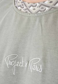 Camicia di cotone verde chiaro con scollatura rotonda, dotata di un colletto interno a motivo e testo ricamato bianco "Project x Paris."