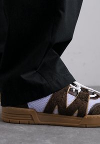 Sneakers cu un superior din suede maro și plasă albă, având accente bej și șireturi rotunde, asortate cu pantaloni negri.