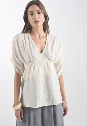 Modella che indossa una leggera e fluida blouse color crema con scollo a V e maniche larghe, abbinata a una gonna grigia, su uno sfondo semplice.