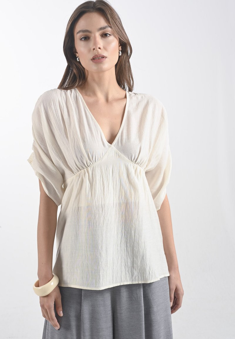 Modella che indossa una leggera e fluida blouse color crema con scollo a V e maniche larghe, abbinata a una gonna grigia, su uno sfondo semplice.