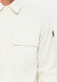 Camicia in corduroy bianco con righe verticali, colletto button-down e una tasca sul petto con un particolare dettaglio in cucitura.