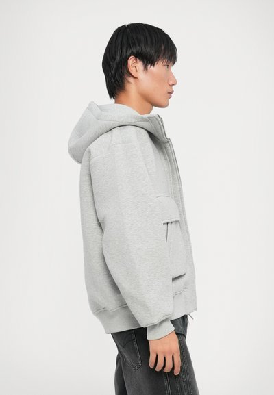 Sweat-shirt gris avec capuche, coupe ample, poignets côtelés et poche kangourou. Texture douce et design simple, porté avec un jean sombre.