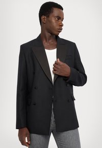 Svart dubbelknäppt kavaj med satinrevers och strukturerade axlar, med en framficka och knappar, ihop med en vit skjorta.