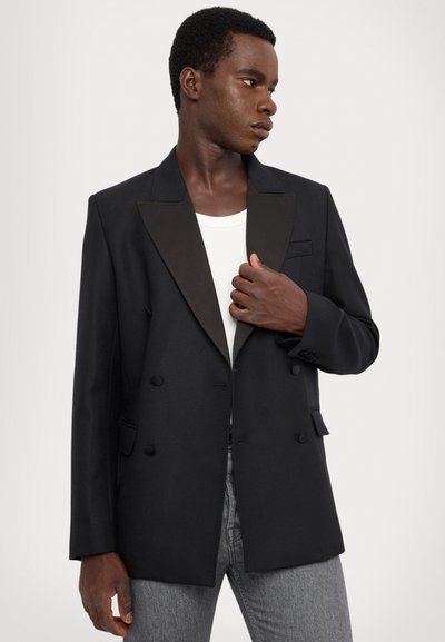 Zwarte dubbelbreasted blazer met satijnen revers en gestructureerde schouders, voorzien van een zak aan de voorkant en knopen, gecombineerd met een wit overhemd.