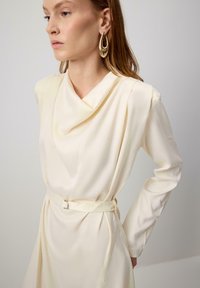 Nais, kes kannab kreemika värvi pika varrukaga kleiti, millel on rippuv kaelus, vöödiga talje ja suured kuldsed ripatsid, vaatab kõrvale.