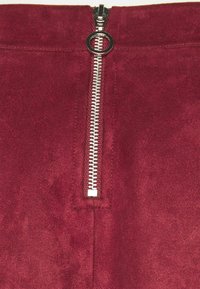 Burgundské kalhoty s měkkou, semišovou texturou. Obsahují stříbrný kovový zip s kulatým táhlem v pase.