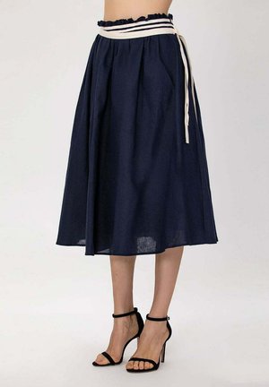 Jupe midi bleu marine avec taille froncée et ceinture rayée blanche, portée avec des talons hauts noirs à bride à la cheville sur fond uni.