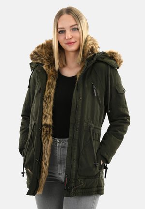Frau in einer olivgrünen Parka mit braunem Kunstpelzbesatz, schwarzem Shirt und grauen Jeans, steht mit den Händen in den Taschen vor einem schlichten Hintergrund.